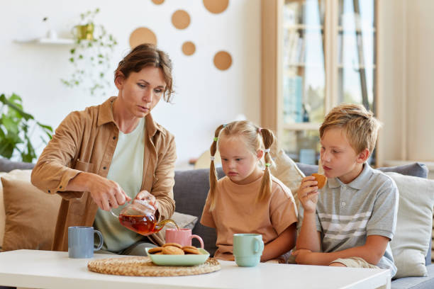 Family Enjoying Tea Portrait of happy family with special need child enjoying tea and desserts at home while sitting together on couch in living room, copy space family dependency mother family with two children stock pictures, royalty-free photos & images