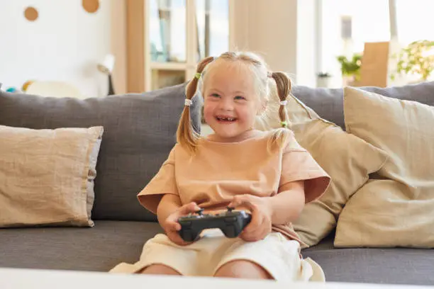 Portrait of cute girl with down syndrome playing video games and laughing happily while sitting on couch in living room, copy space