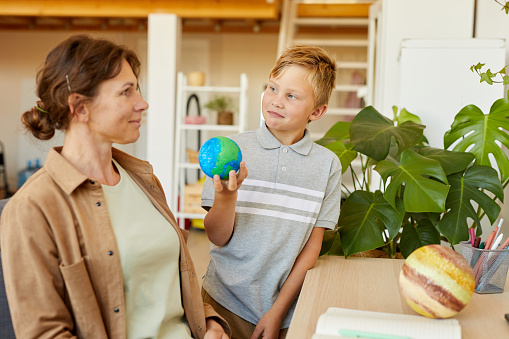 Portrait of blonde teenage boy holding planet model while studying at home with mother , copy space