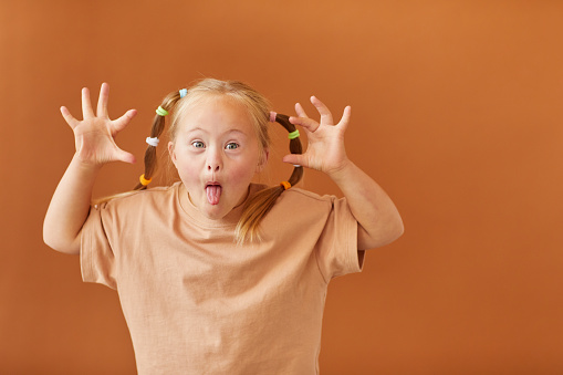 Waist up portrait of cute girl with down syndrome making faces at camera while standing against plain brown background in studio, copy space