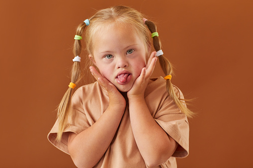 Waist up portrait of cute girl with down syndrome grimacing at camera while standing against plain brown background in studio, copy space