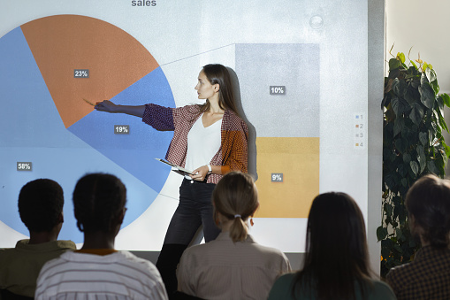 Side view portrait of young woman pointing at data chart with statistics while giving presentation during business conference, copy space