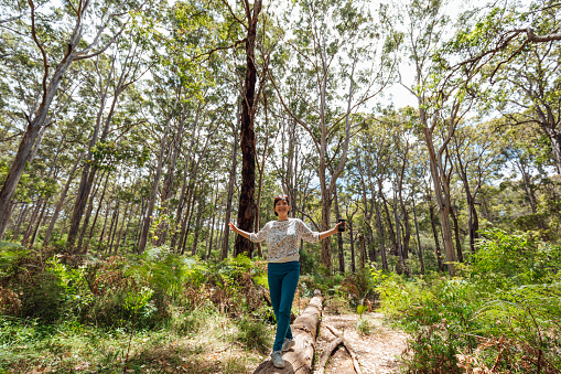 A shot of a mature asian woman walking and balancing on a log in a forest. She is wearing casual clothing and is surrounded by large trees and shrubbery.