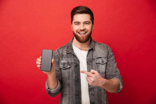 istock Image of young brunette man pointing finger on smartphone and smiling on camera, isolated over red background 1275860244