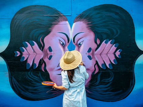 Female mural artist creating art on the wall exterior in public park in the city.