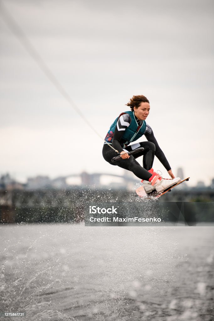 woman in wetsuit holding rope and professionally jumping over water on wakeboard woman in wetsuit holding rope in her hand and professionally jumping over splashing water on wakeboard Wakeboarding Stock Photo
