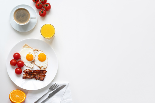American breakfast with two eggs, bacon, tomatoes, orange, coffee and orange juice on the white table. Top view. Copy space.