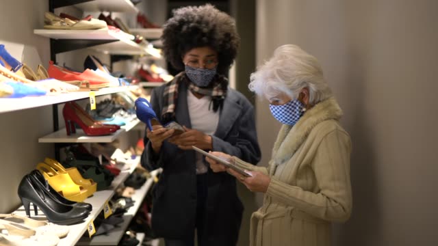 Senior woman wearing face mask being helped by a saleswoman while shopping for shoes in a thrift store