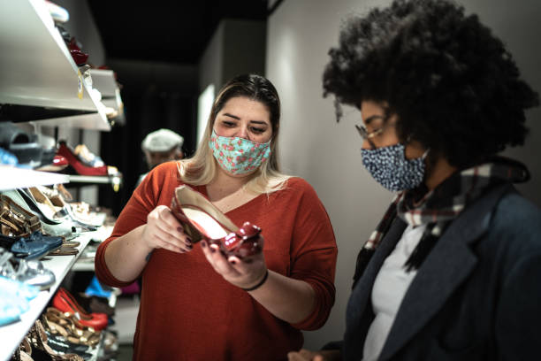 giovane donna che indossa la maschera facciale aiutata da una commessa mentre fa shopping per le scarpe in un negozio di parsimonia - shoe store sales clerk customer foto e immagini stock