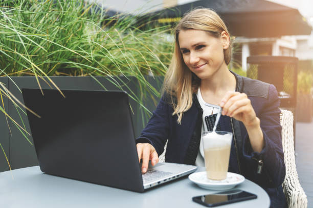 カフェの屋外テラスでコーヒーラテを飲んで、ラップトップで作業する若い女性。フリーランスのリモートワーク - furniture internet adult blond hair ストックフォトと画像