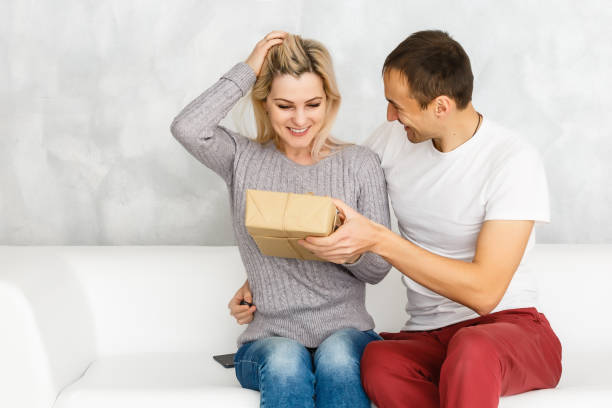 istock Handsome young man is giving a present to his lovely girlfriend while they are sitting on couch at home 1275872797