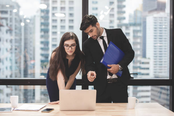 Businessman and businesswoman using a laptop together while standing in front of office building windows overlooking the city. Two confident businesspeople using a laptop working at a table in a modern office hombre de negocios y mujer de negocios que utilizan un ordenador portátil juntos mientras están de pie frente a las ventanas del edificio de oficinas con vistas a la ciudad. dos empresarios seguros que usan un portátil trabajando en una mesa en una ofic