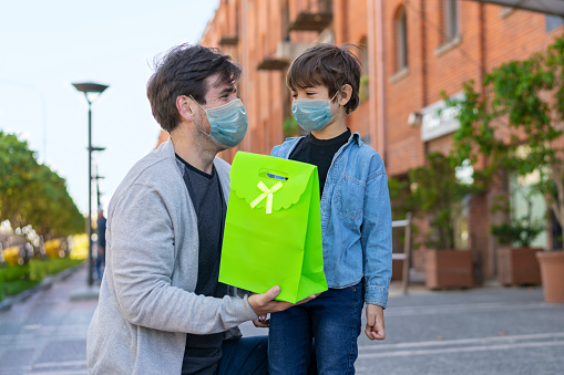 Loving son hugging daddy while he holds a gift for father's day both looking at each other wearing protective facemasks - Pandemic lifestyles