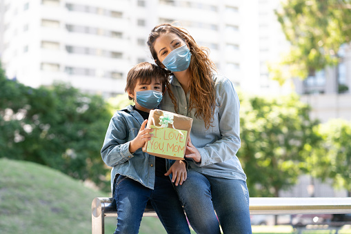 Portrait of loving mom and son at the park hugging each other holding a mother's day gift looking at camera wearing protective facemasks - Pandemic lifestyles