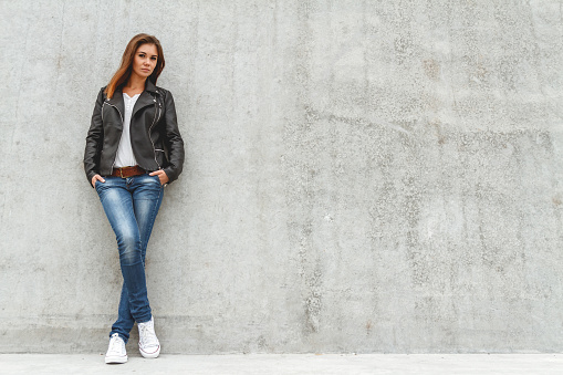 Portrait of a beautiful girl in jeans and a black leather jacket from the concrete wall