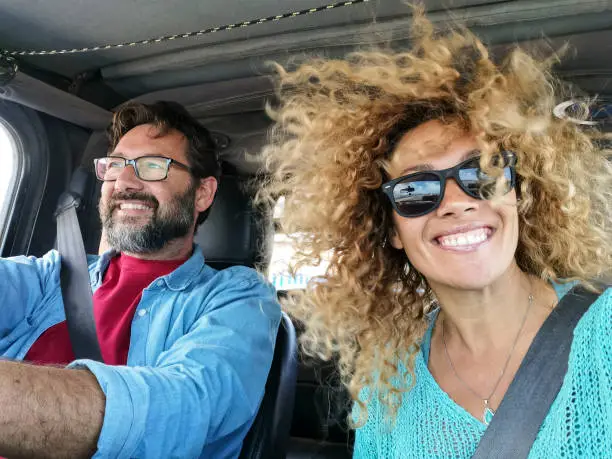 Photo of Cheerful couple drive and enjoy the trip inside a car with wind in the hair - beautiful young woman smile at the camera - people travel on vehicle concept