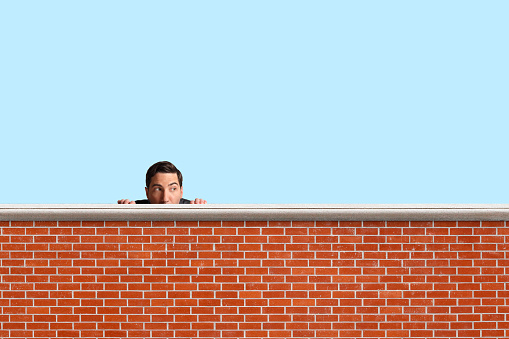 A businessman peers over the top of a brick wall with blue sky in the background.