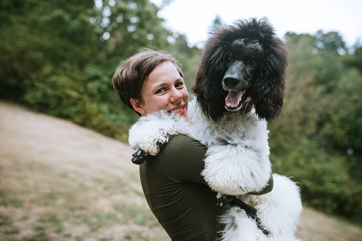 A Caucasian young adult woman with her standard poodle at a park, enjoying the company of her beloved pet.  She holds the large dog in a loving embrace.  Shot in Washington State, USA.