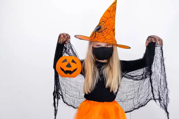 Photo of Portrait blonde girl in witch costume wearing face mask with candy bucket pumpkin preparing for Halloween on white studio background. copyspace