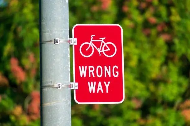 Red bicycle wrong way sign on street pole placed facing wrong-way bicycle traffic. Blurred trees background