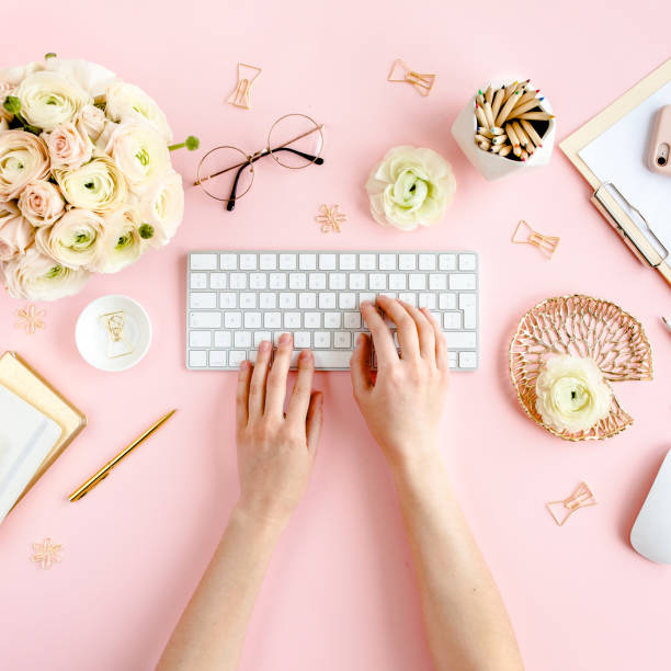 istock Stylized, pink women's home office desk. Workspace with female hands, computer, bouquet ranunculus and roses, clipboard on pink background. Flat lay 1276140797
