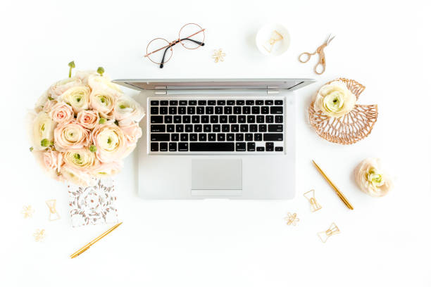 istock Stylized women's home office desk. Workspace with laptop, computer, bouquet ranunculus and roses, clipboard on white background. Flat lay. Top view 1276152106