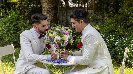 Young gay couple in white tuxedo enjoys lovely moment with each other during wedding ceremony at home. Two gays sharing their happy feeling at a garden in a house with love and care
