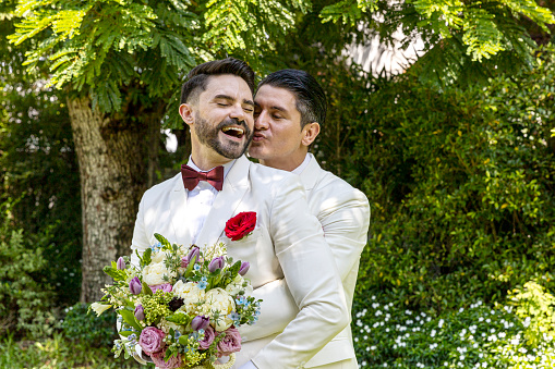 Young gay couple in white tuxedo holding each other during wedding ceremony at home. Two gays sharing their happy feeling at a garden in a house with love and care