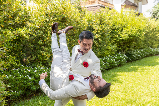 Two gays in white suit sharing their happy feeling in a garden at home during wedding ceremony. One gay is lifting his partner with care and love