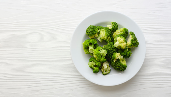 Colorful contrasting freshly steamed bright green healthy organic broccoli spears on a white ceramic plate sitting on an abstract white wood table background with lots of grain character, shot from directly above with good copy space on the left of the image.