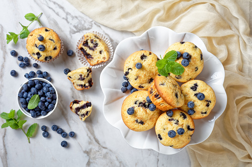 Blueberry muffins baked with fresh blueberries on a white cake stand with fresh mint on top on a white wooden background, top view, close-up
