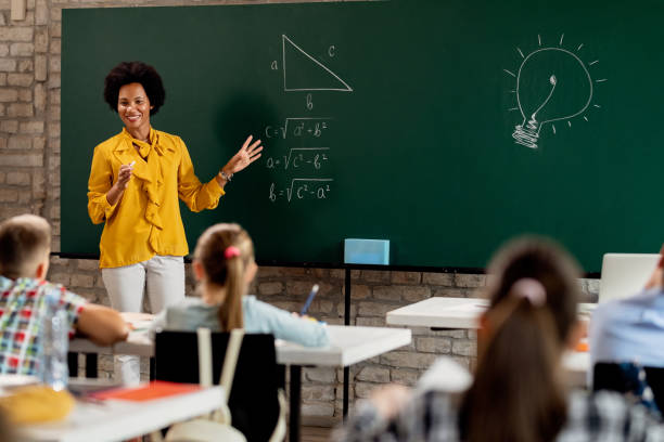 Happy African American math teacher explaining lecture on blackboard in the classroom. Happy black elementary school teacher talking to her students and explaining them mathematics on blackboard in the classroom. math teacher stock pictures, royalty-free photos & images