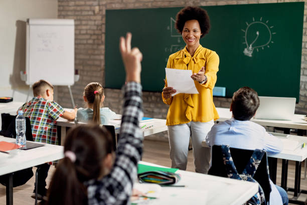 Please, tell us a correct answer! Happy African American teacher holding a class and pointing at student with raised arm who wants to answer the question. elementary student pointing stock pictures, royalty-free photos & images