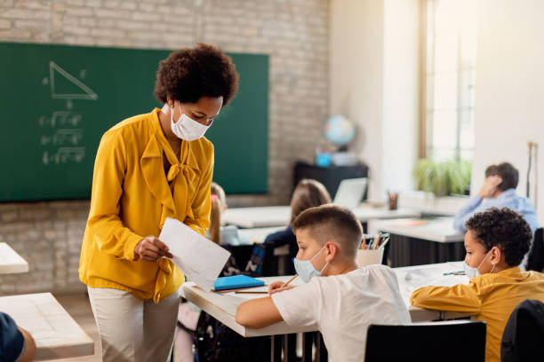 Black teacher and elementary student with face masks examining test result in the classroom. African American explaining test results to a schoolboy during a class. They are wearing protective face masks due to COVID-19 pandemic. elementary school building photos stock pictures, royalty-free photos & images