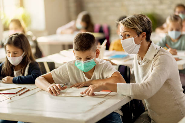 istock Female teacher and elementary student wearing protective face mask during a class in the classroom. 1276372662