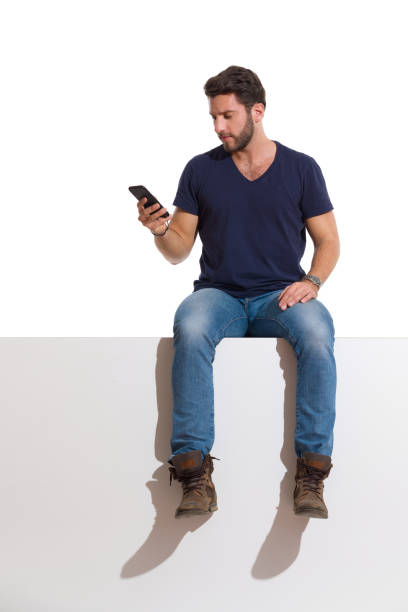 Handsome Man Is Sitting Relaxed On A Top, Holding Telephone And Reading Focused man in boots, jeans and unbuttoned lumberjack shirt is sitting on a top, holding telephone and reading. Full length studio shot isolated on white. man beard plaid shirt stock pictures, royalty-free photos & images