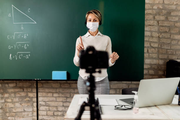 Teacher with face mask giving math lesson online from the classroom. Female teacher standing in front of blackboard and explaining math online during coronavirus pandemic. teacher classroom child education stock pictures, royalty-free photos & images