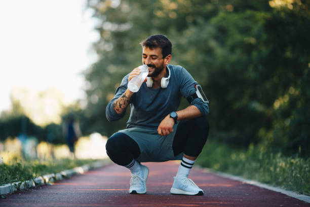 młody atrakcyjny uśmiechnięty sportowiec kucający na bieżni i wody pitnej. - drinking men water bottle zdjęcia i obrazy z banku zdjęć