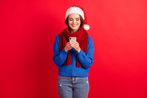 Photo of positive girl in santa claus cap use smartphone typing christmas x-mas newyear congratulations wear blue knitted sweater, denim jeans isolated over bright shine color background