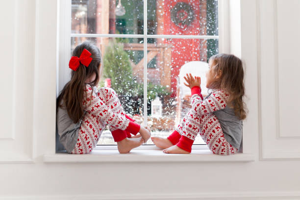 zwei niedliche mädchen im pyjama sitzen und schauen aus dem fenster bei schneebedecktem wetter. weihnachtsmomente mit kindern zu hause - 3 4 months stock-fotos und bilder