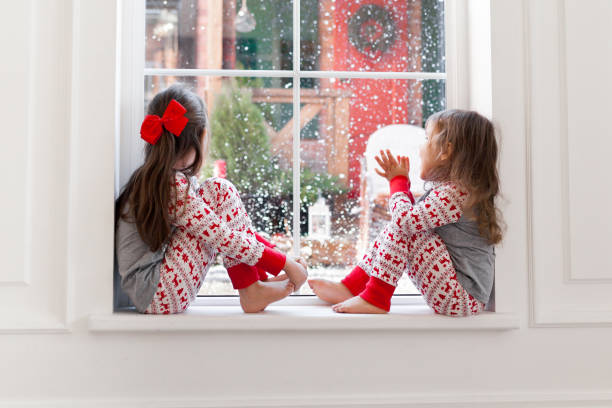 due ragazze carine in pigiama seduti e guardando fuori dalla finestra a tempo nevoso. momenti di natale con i bambini a casa - christmas home foto e immagini stock