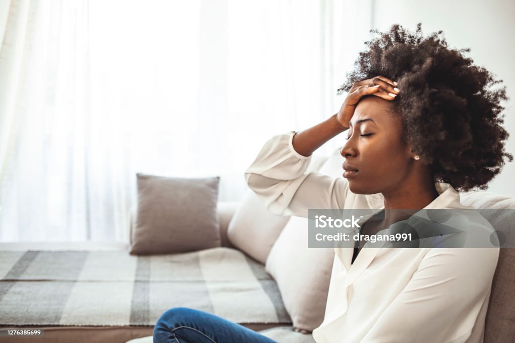 Shot of a young woman suffering from a headache. Lonely sad woman deep in thoughts sitting daydreaming or waiting for someone in the living room with a serious expression, she is pensive and suffering from insomnia sitting on couch Emotional Stress Stock Photo
