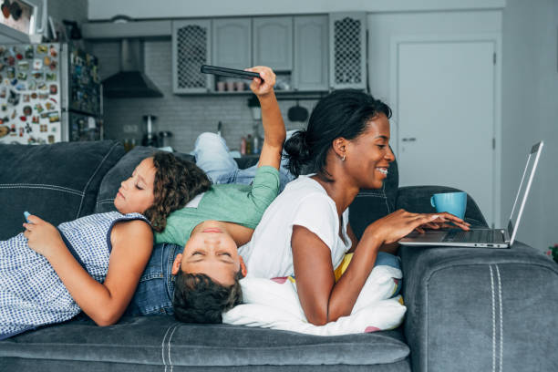 Digital family Mixed race family sitting on the sofa at home and using digital devices. Smiling african-american mother using laptop while her two children using smartphones. family dependency mother family with two children stock pictures, royalty-free photos & images