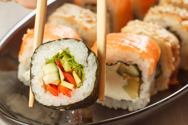 istock Two chopsticks holding Hosomaki with vegetables and different sushi rolls behind 1276389383