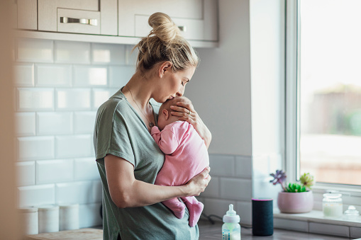 A young woman standing in her kitchen, she is having a tender moment with her newborn baby girl.
