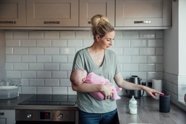 Turning the Music Down A young woman standing in her kitchen, holding her newborn daughter in her arms while adjusting the volume on her smart speaker. virtual assistant stock pictures, royalty-free photos & images
