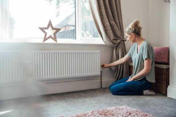 Reducing Energy Consumption Side view of a young woman kneeling on the living room floor, adjusting the thermostat on the radiator. radiator heater stock pictures, royalty-free photos & images