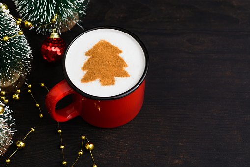 Christmas red cup of coffee with fir tree pattern. New Year beverage over dark wooden background with copy space. Mag of cappuccino with art in shape of Christmas tree and holiday decorations