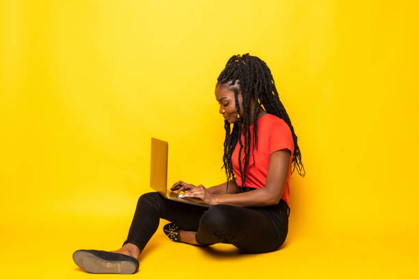 istock Happy afro woman using laptop, talking on phone while sitting on floor over yellow background 1276396239