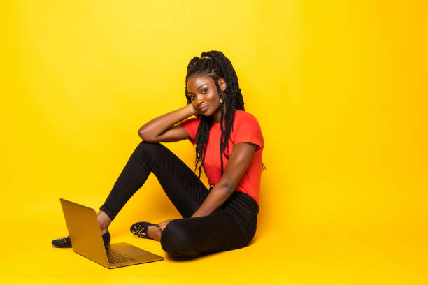istock Happy afro woman using laptop, talking on phone while sitting on floor over yellow background 1276399677